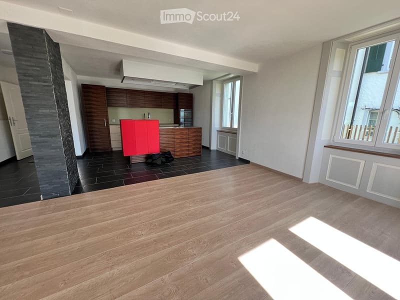 Open-plan living and kitchen area with wooden flooring, modern kitchen cabinets, a red accent cabinet, and a sink with a faucet. The living area has large windows and a wooden floor.