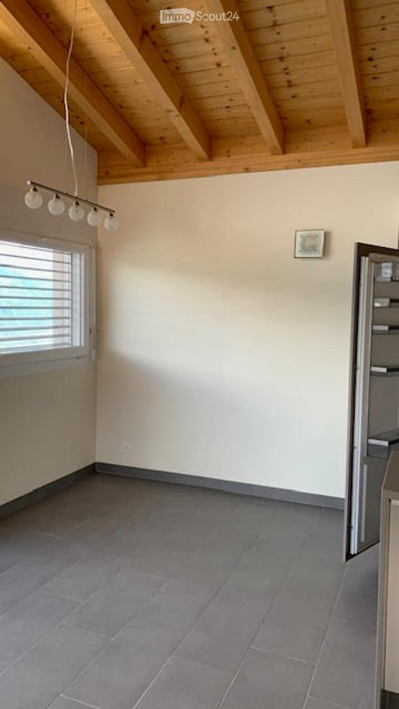 Empty room with wooden ceiling beams, grey tiled floor, large window with blinds, and a partially visible kitchen area with a stainless steel appliance.
