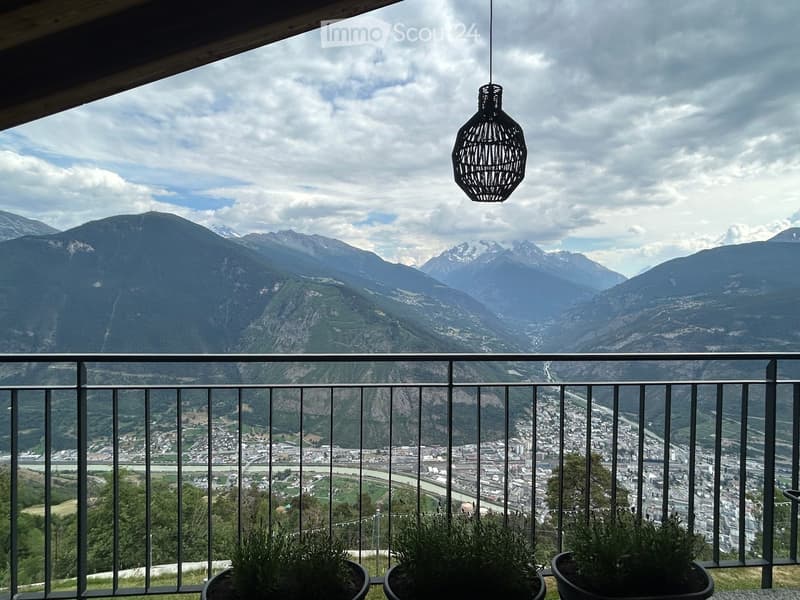Balcony with metal railing, hanging woven pendant light, and potted plants, overlooking a town and mountain range.