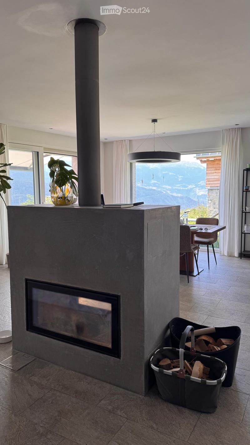 Modern living room with a large fireplace, pendant light, dining area with chairs and table, and a vase with plants on the mantel. Offers a scenic mountain view through large windows.