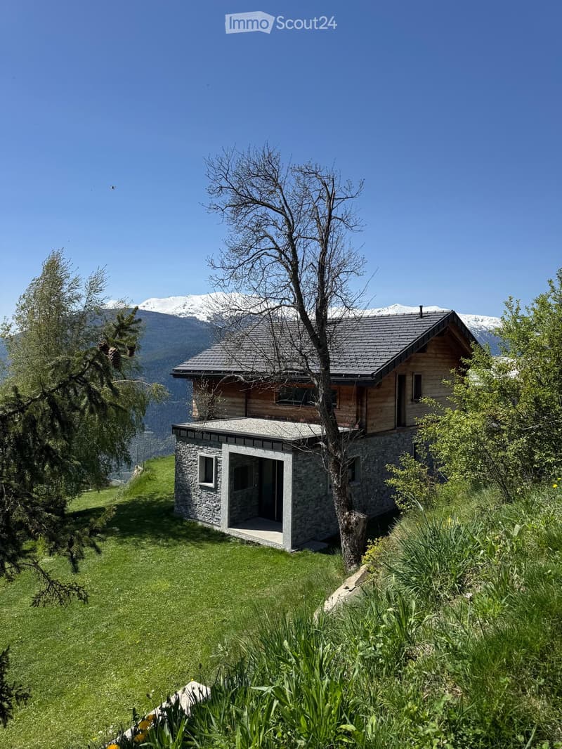 A two-story wooden house with a stone base, situated on a grassy slope with a view of snow-capped mountains in the background.