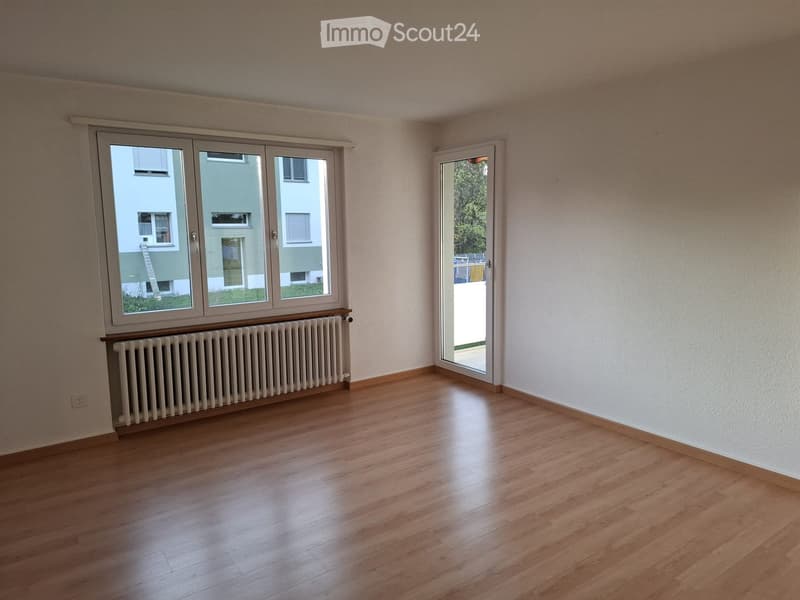 Empty room with wooden flooring, large windows, radiator, and a sliding door leading outside.
