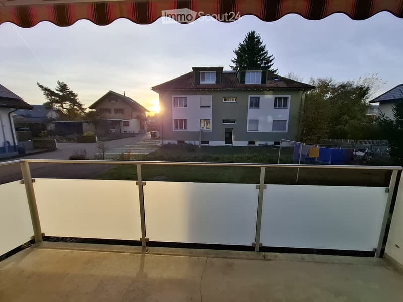 Balcony with metal railing, overlooking a residential area with houses, trees, and a sunset view.