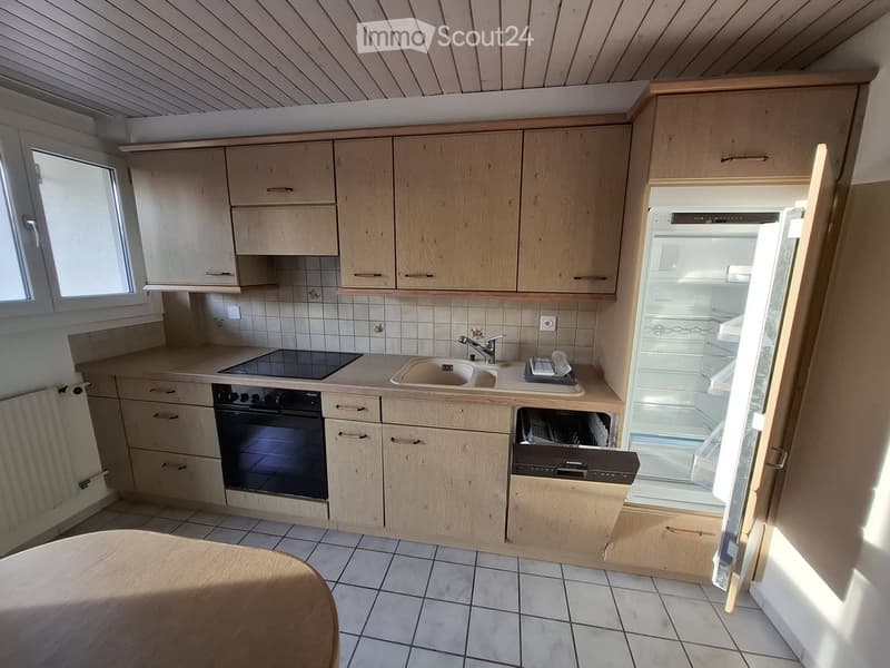 Modern kitchen with light wooden cabinets, tiled walls, and appliances including a sink, oven, dishwasher, and refrigerator. A round wooden table is partially visible in the foreground.