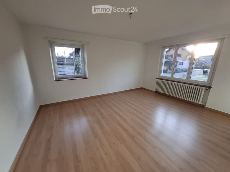 Empty room with wooden flooring, white walls, and two windows offering a view of the outside. A radiator is visible under one window.
