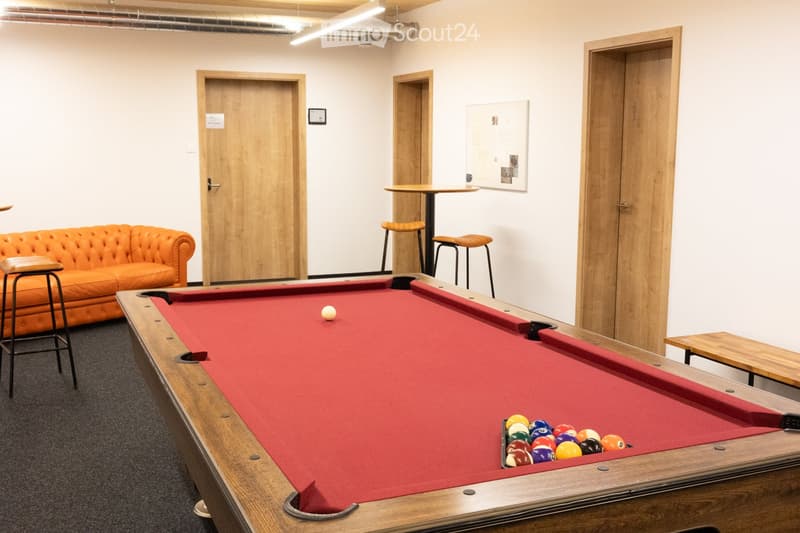 A modern, well-lit room with a pool table, orange sofa, and wooden stools. The room features white walls, wooden doors, and a small table with chairs.