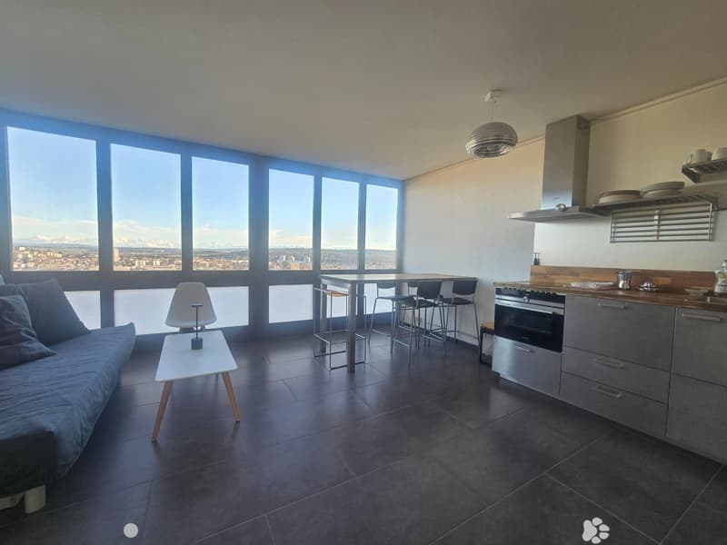 Modern open-plan living and dining area with a sofa, small table, dining table with chairs, and a kitchen area with an oven and cabinets. Large windows offer a scenic view.