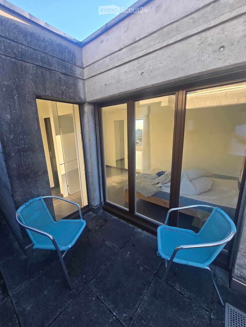 Balcony with two blue chairs, large windows showing a bedroom with a bed and pillows.
