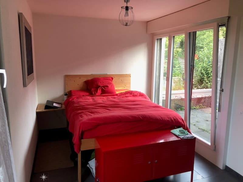 Cozy bedroom with a wooden bed frame, red bedding, a nightstand, and a red cabinet. Large windows offer a view of greenery outside.