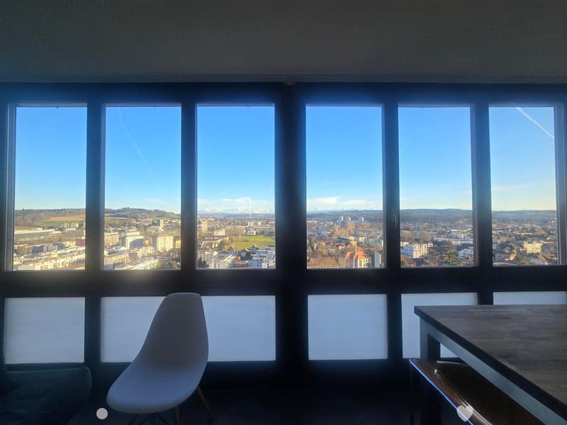 A modern, minimalist room with large windows offering a panoramic city view. The room features a sleek white chair and a wooden table.