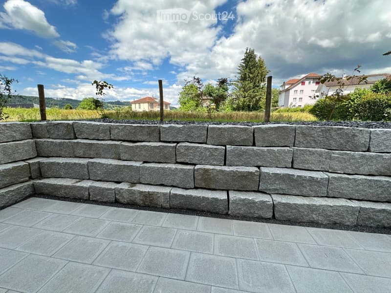 A stone retaining wall with a paved area in front, surrounded by greenery and distant buildings.