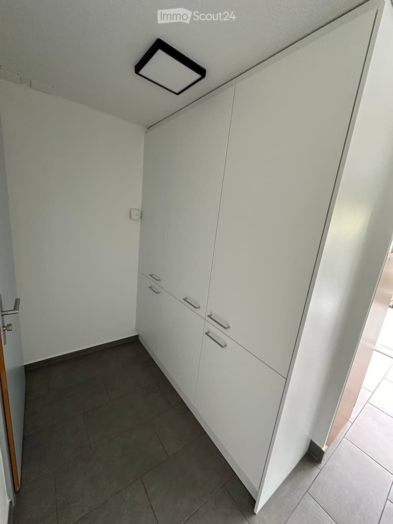 A narrow hallway with white walls, dark tiled floor, white built-in cabinets with silver handles, and a square ceiling light fixture.