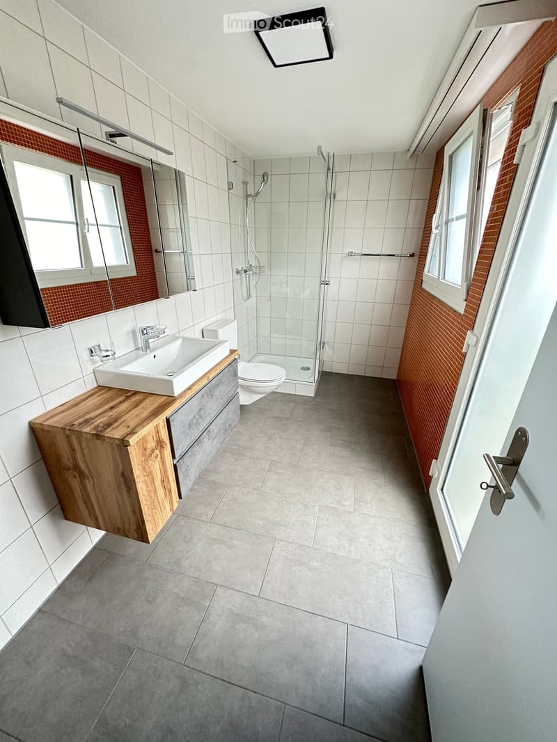 Modern bathroom with white tiles, grey floor, wooden vanity cabinet, sink, toilet, and glass-enclosed shower. Features large windows and a sleek design.