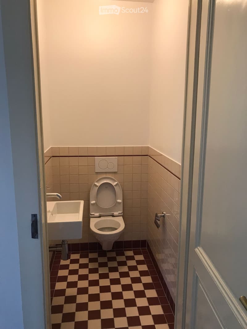 Small bathroom with a toilet, sink, and tiled walls. The floor has a checkered pattern in brown and beige tiles.