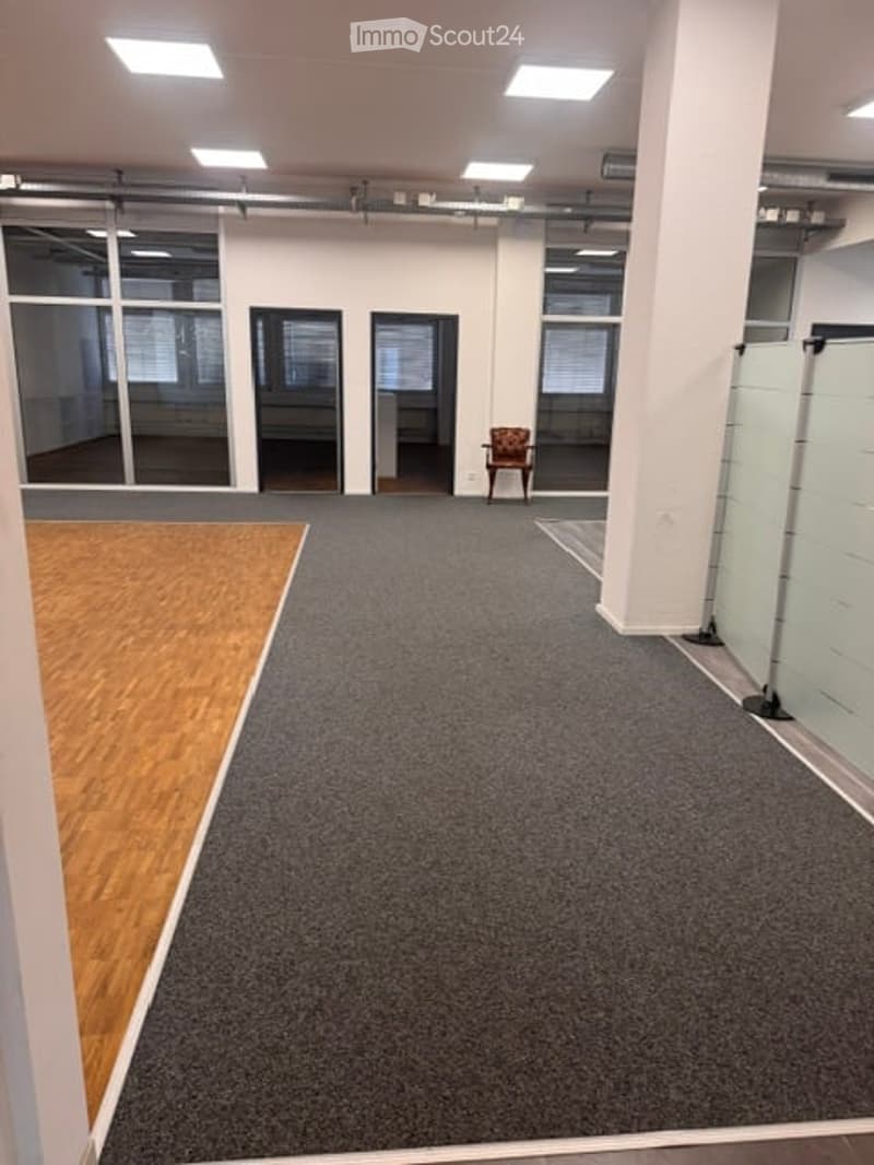 Open-plan unfurnished room with grey carpeted floor, large windows, and a single wooden chair near the back wall.