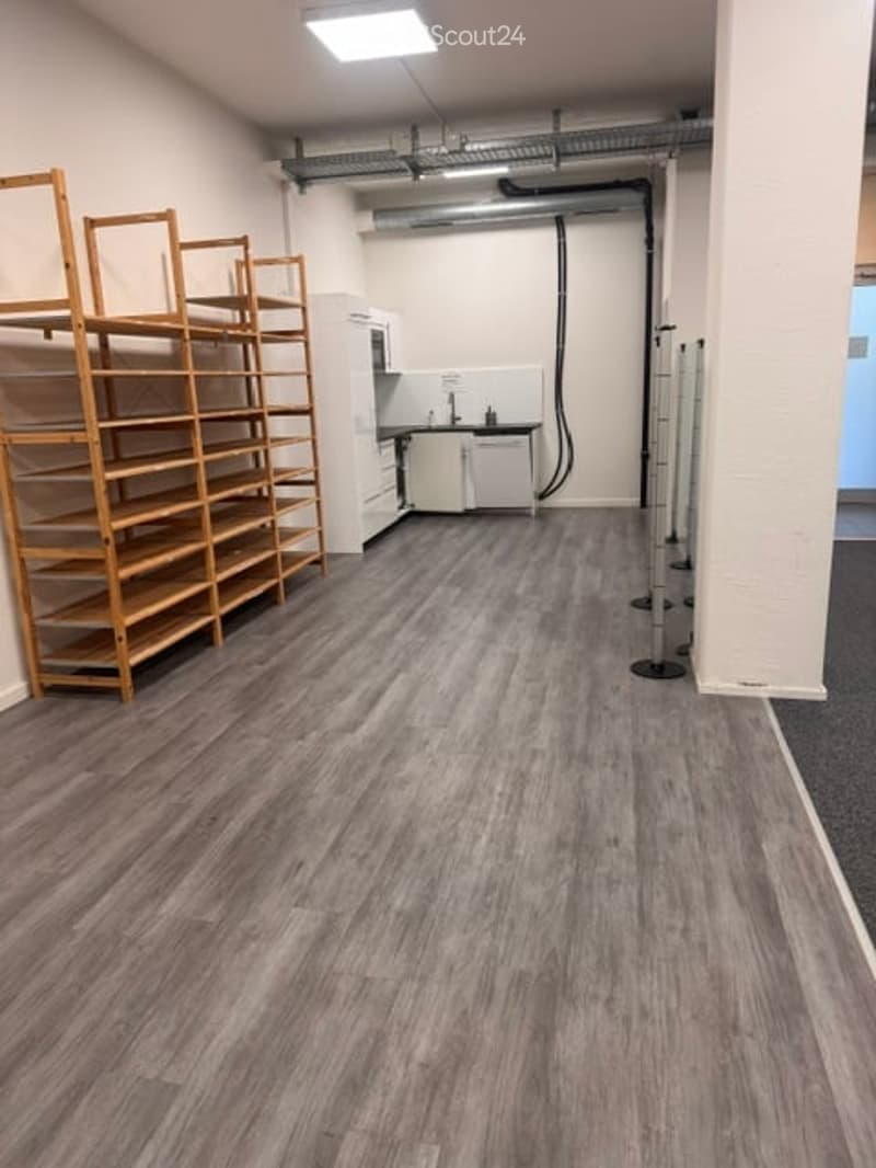 The room features a wooden shelving unit on the left, a small kitchen area with cabinets and a sink, and a grey laminate floor. The space is well-lit with a ceiling light.