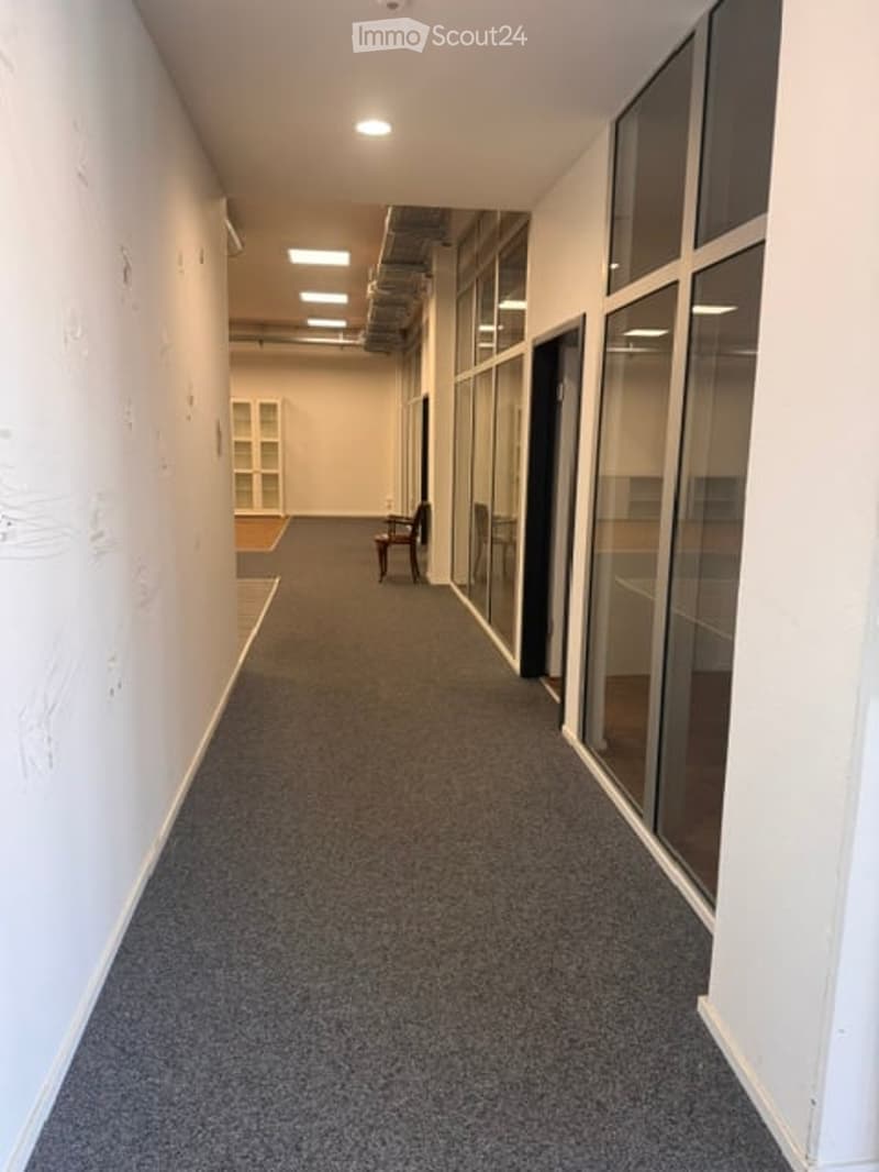 Clean, modern hallway with grey carpeting, white walls, and large glass windows on one side. A single chair is placed against the wall near the end of the corridor.
