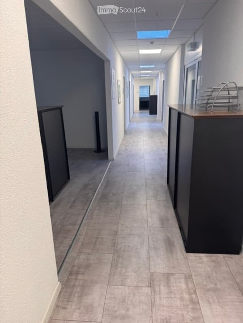 Clean, modern hallway with gray tiled flooring, white walls, and recessed lighting. Features a reception area with a dark wooden counter and a small seating area visible at the end.