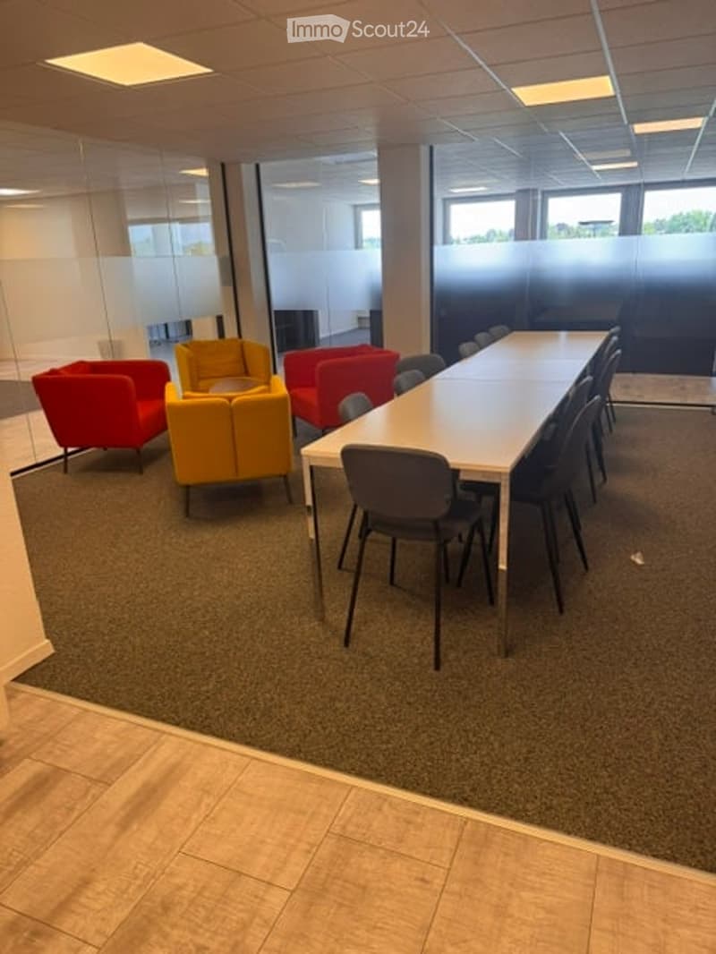 A modern meeting or break room with a long white table surrounded by black chairs, colorful sofas, and large windows with blinds.