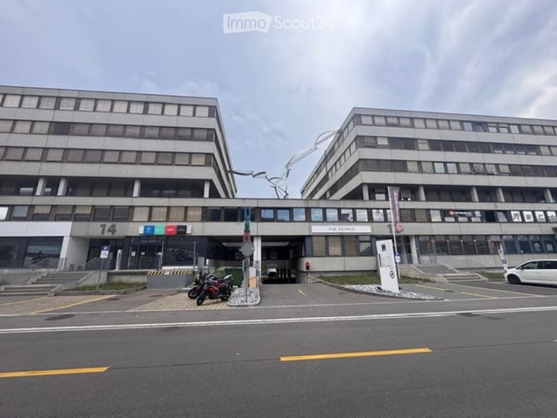 Modern multi-story building with a symmetrical facade, numbered '14', and a small entrance area with a few parked motorcycles and a car.