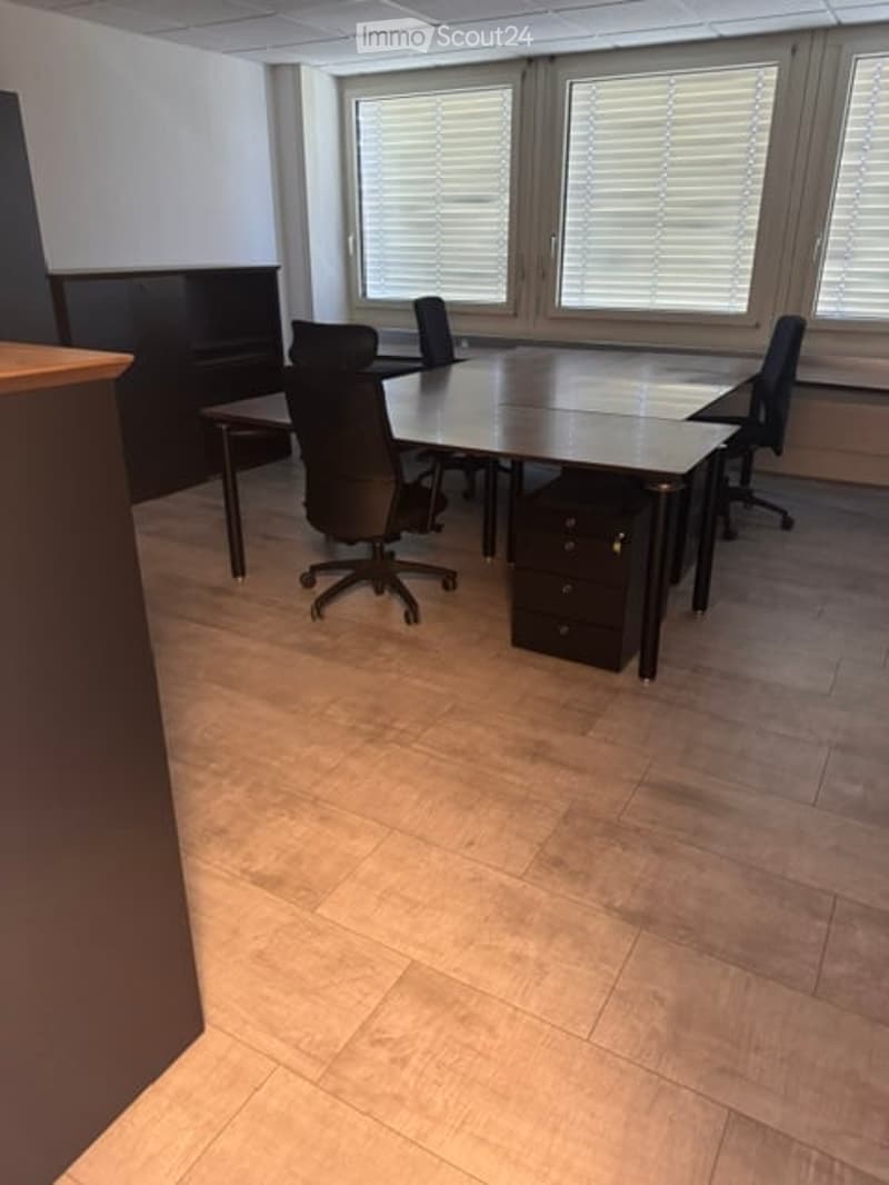 Office space with a large conference table, several office chairs, and a cabinet. The room has large windows with blinds and a tiled floor.