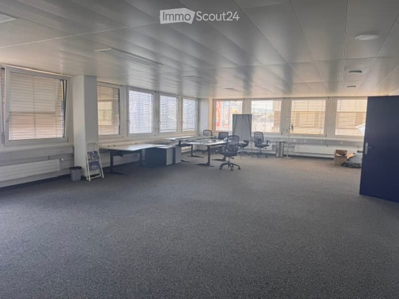 Office space with multiple desks, chairs, and a large window with blinds. The room has a carpeted floor and white walls.