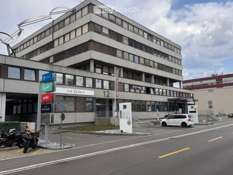 Multi-story commercial building with a modern facade, signage for various businesses, and a parking area with motorcycles and a car.
