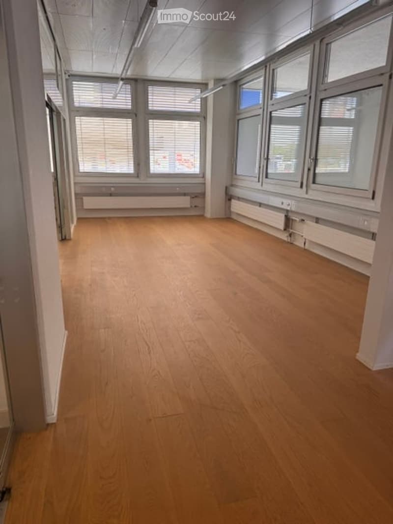 Empty room with wooden flooring, large windows with blinds, and white walls.
