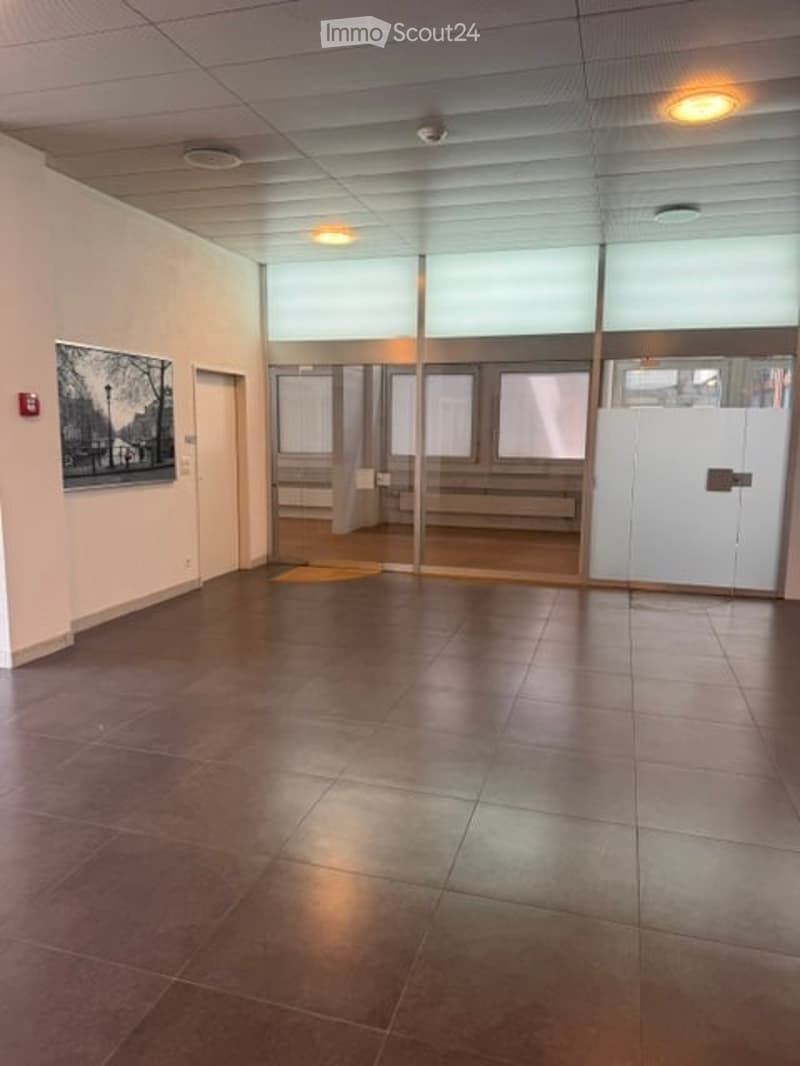 Empty room with tiled floor, white walls, large windows, and a glass partition leading to another area.