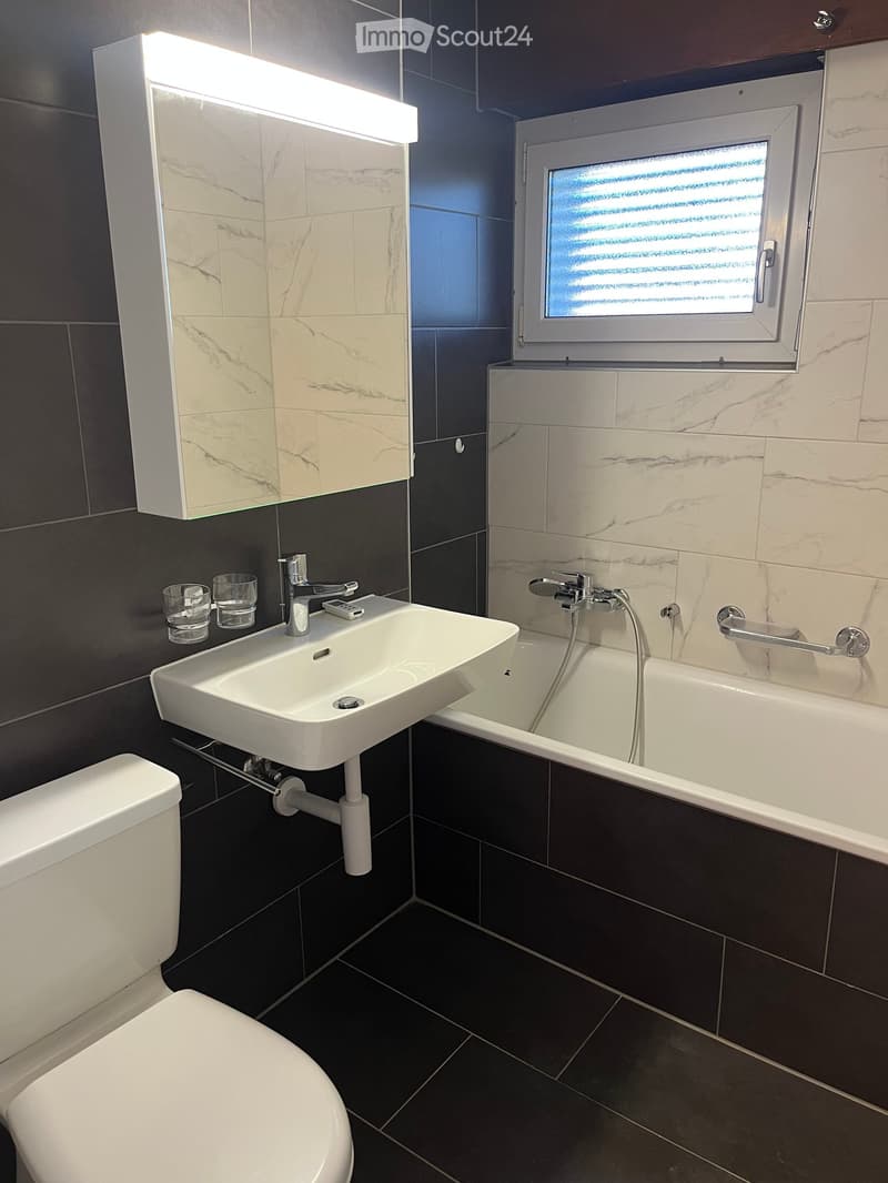Modern bathroom with a white sink, toilet, bathtub, and a large mirror with built-in lighting. The walls are tiled in a combination of dark and light marble patterns, and the floor is dark tiled.