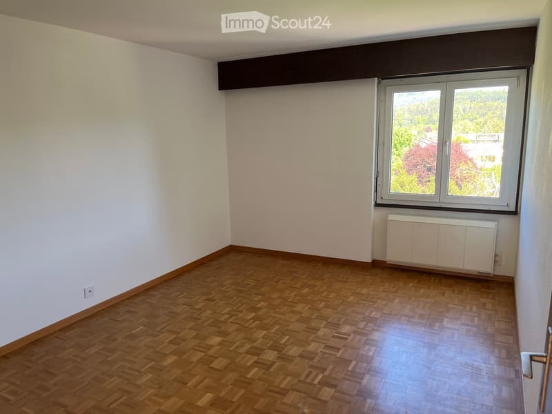 Empty room with wooden flooring, white walls, and a large window with a view of greenery. A radiator is visible below the window.