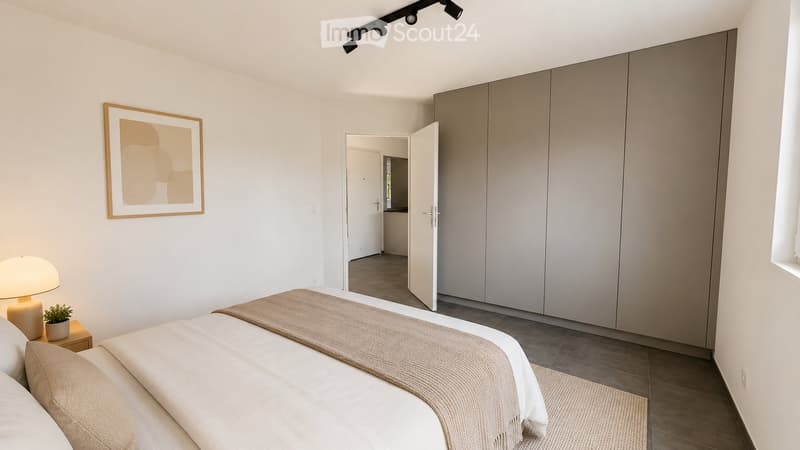 Modern bedroom with a neatly made bed, nightstand with a lamp, small plant, and a large wardrobe. A picture frame hangs on the wall, and an open doorway leads to another room.