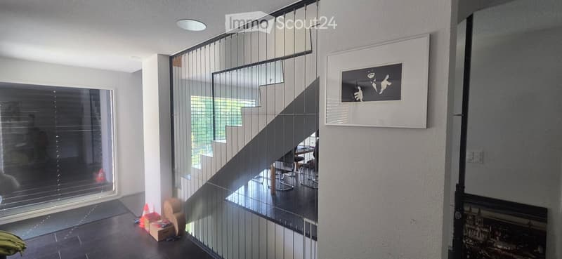 Modern hallway with glass staircase, large windows, artwork on walls, and a view of a lower level room with chairs and a table.