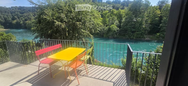 Balcony overlooking a river with a yellow table and colorful chairs.