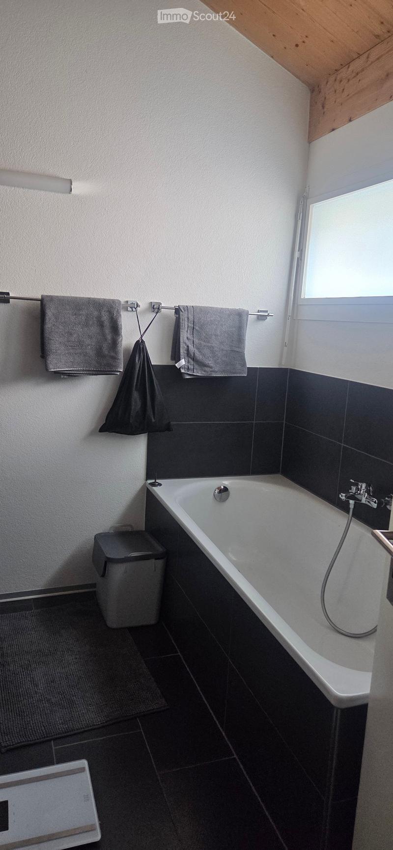 Modern bathroom with a white bathtub, dark tiled walls and floor, towel rack with towels, and a trash bin.