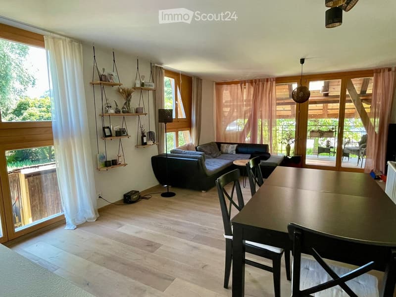 Open-plan living and dining area with wooden flooring, large windows, and a balcony. Features a sofa, dining table with chairs, shelves, and floor lamp.
