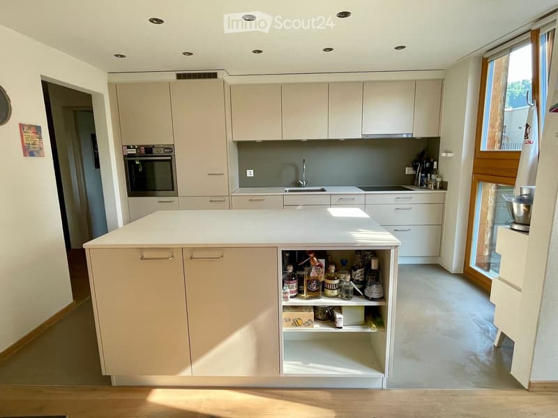 Modern kitchen with white cabinets, central island, built-in oven, sink, and various appliances. Shelves hold wine bottles and other items.