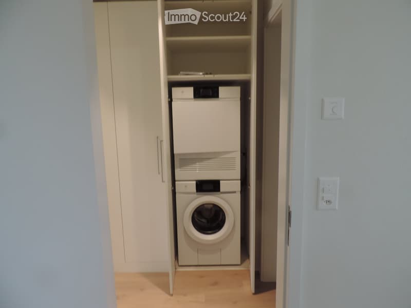A small, well-lit room with a wooden floor and white walls, featuring a built-in closet with a washing machine and a dishwasher inside.