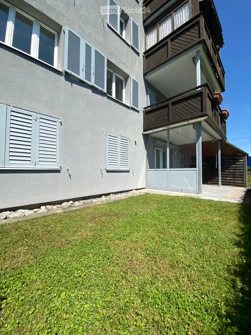 The front of an apartment building with windows, shutters, and balconies on the upper floors, with a grassy area in front.