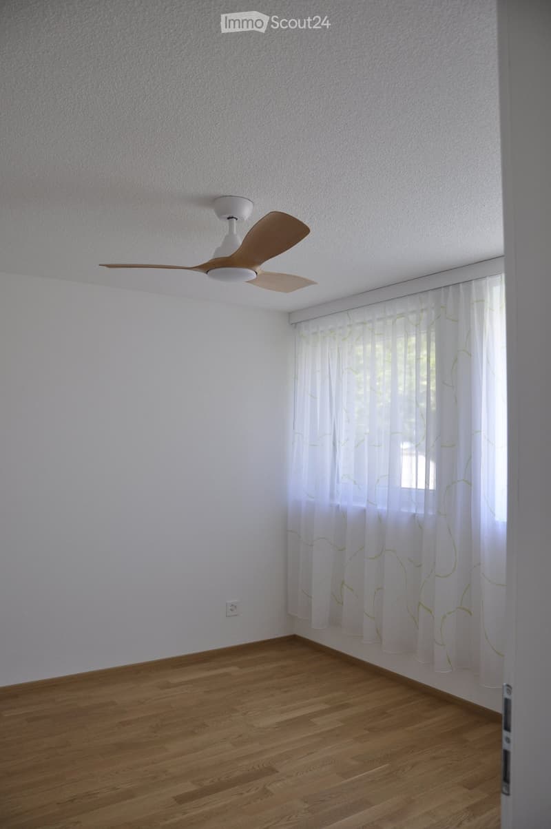 Empty room with light-colored walls, wooden flooring, and a ceiling fan with wooden blades. A window with white curtains is visible on one wall.