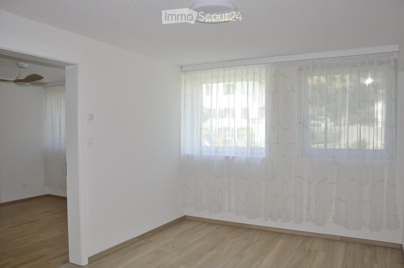 Empty room with white walls, wooden flooring, and large windows covered with sheer curtains. A ceiling fan is visible in the adjacent room.