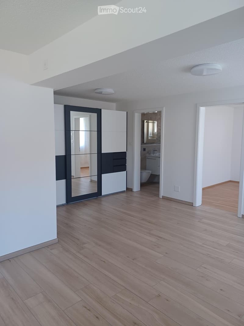 Empty room with wooden flooring, white walls, and a large mirrored wardrobe. A bathroom is visible through an open doorway.