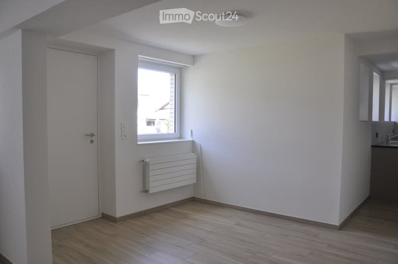 Empty room with wooden flooring, white walls, a window, a radiator, and a door. No furniture or equipment is present.