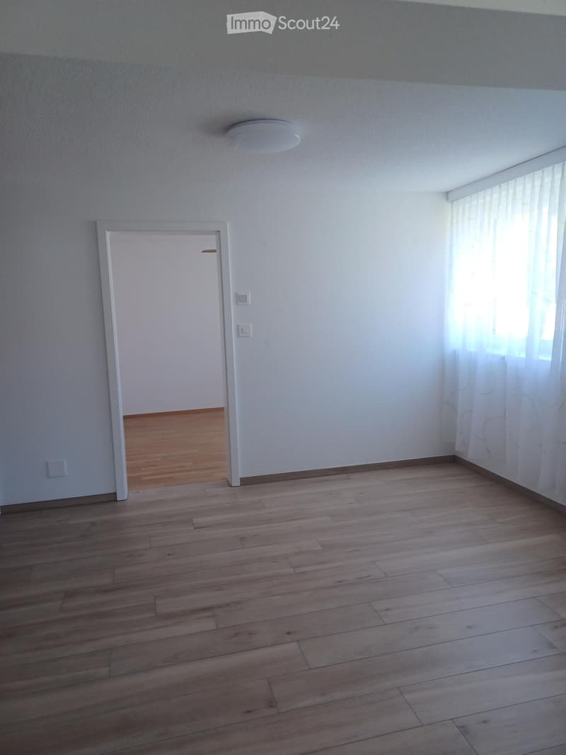 Empty room with wooden flooring, white walls, and a large window with curtains. A door leads to another room.