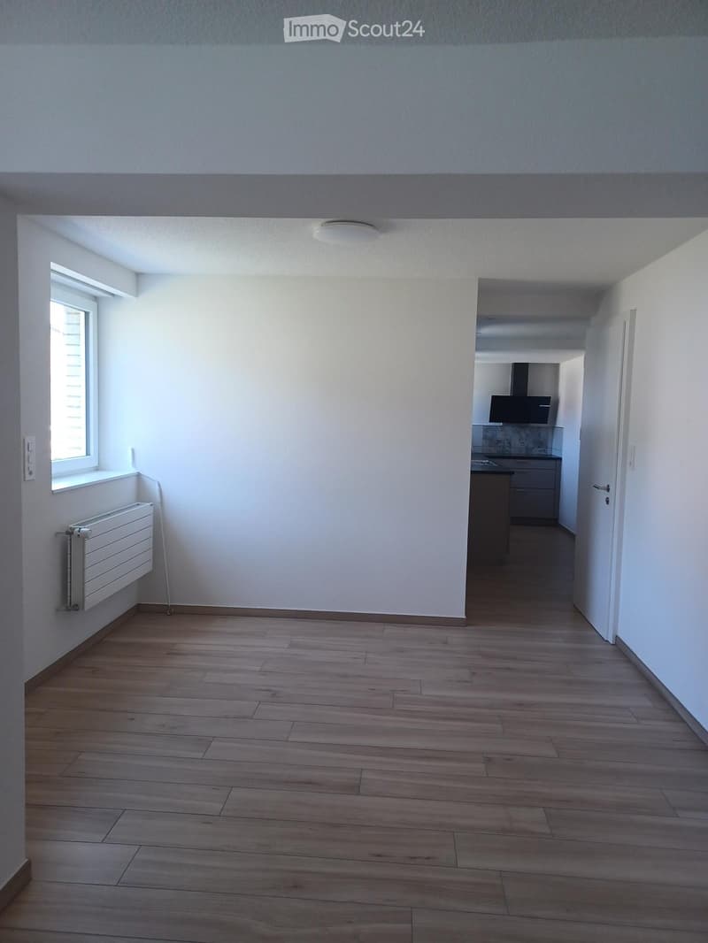 Empty room with wooden flooring, white walls, and a window with blinds. A radiator is mounted on the wall near the window, and a doorway leads to another room.