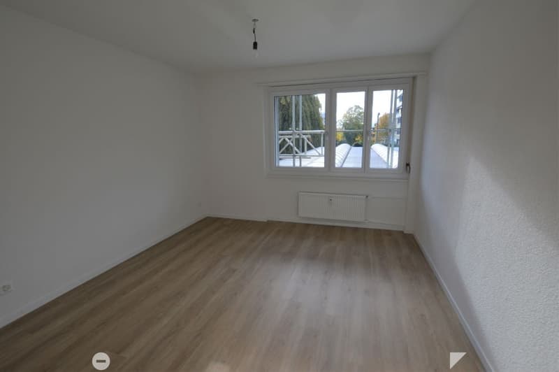 Empty room with wooden flooring, white walls, and a large window offering a view of a balcony area.