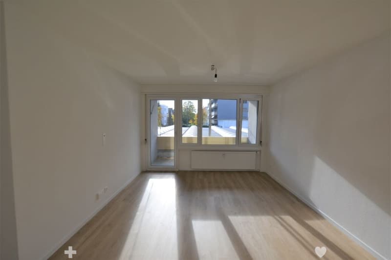 Empty room with wooden flooring, white walls, large window with glass door leading to a balcony, radiator, and visible electrical outlets.