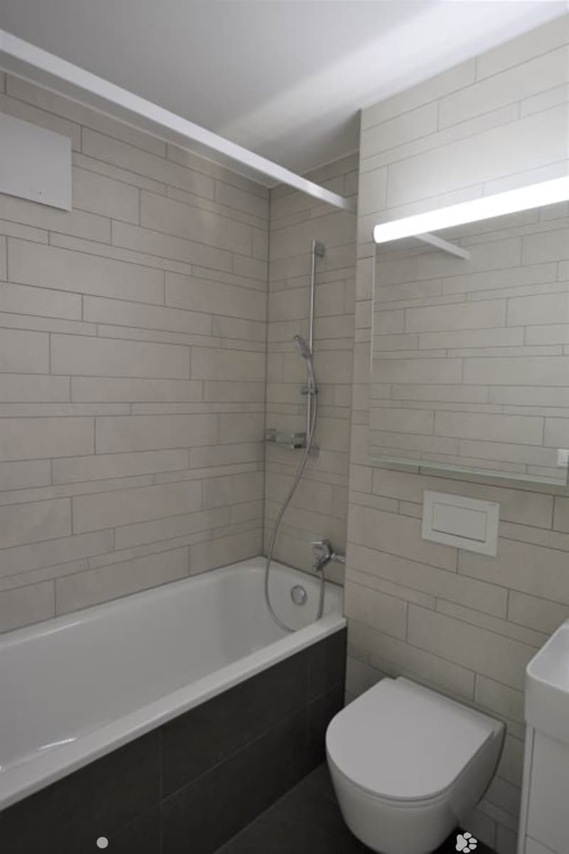 Modern bathroom with a bathtub, toilet, shower, and a mirror with a shelf above it. The walls are tiled in a light grey color, and the floor is dark.