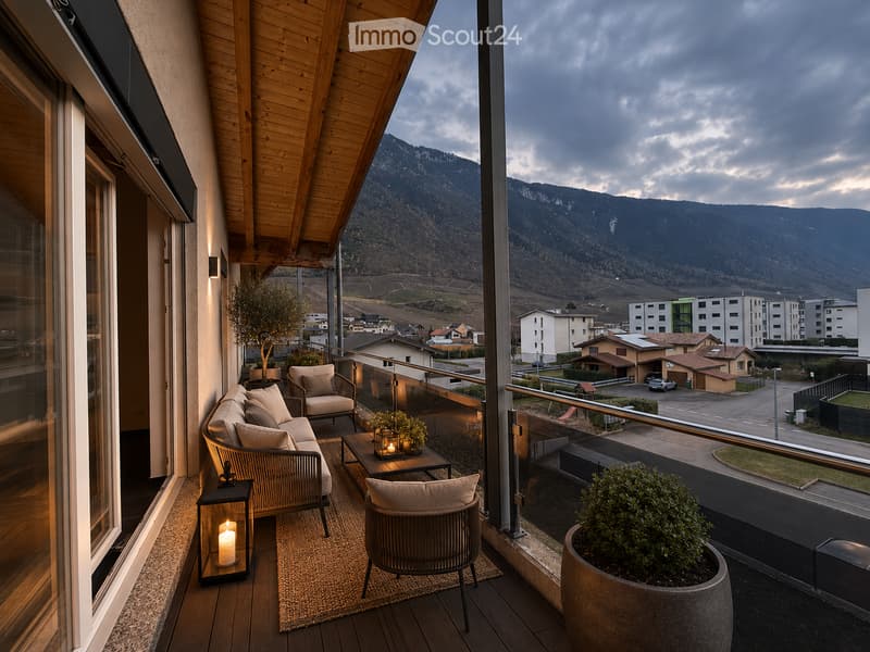 Balcony with wooden decking, sofa, chairs, small table, lantern, and potted plants, overlooking a town and mountains.