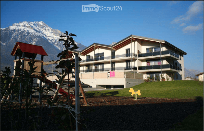 A modern two-story residential building with balconies and a garage, set against a backdrop of snow-capped mountains.